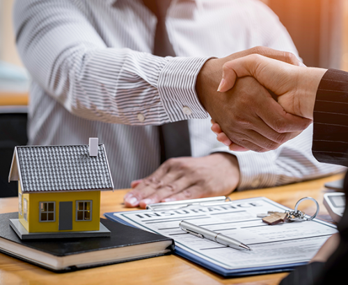 Real estate transaction: handshake agreement with documents, house model, and Zen Home Realty branding on table.